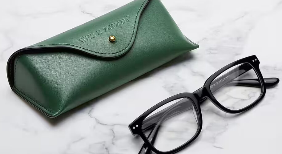 Black-rimmed eyeglasses with green Timo x Zaneti embossed leather case on a marble surface.