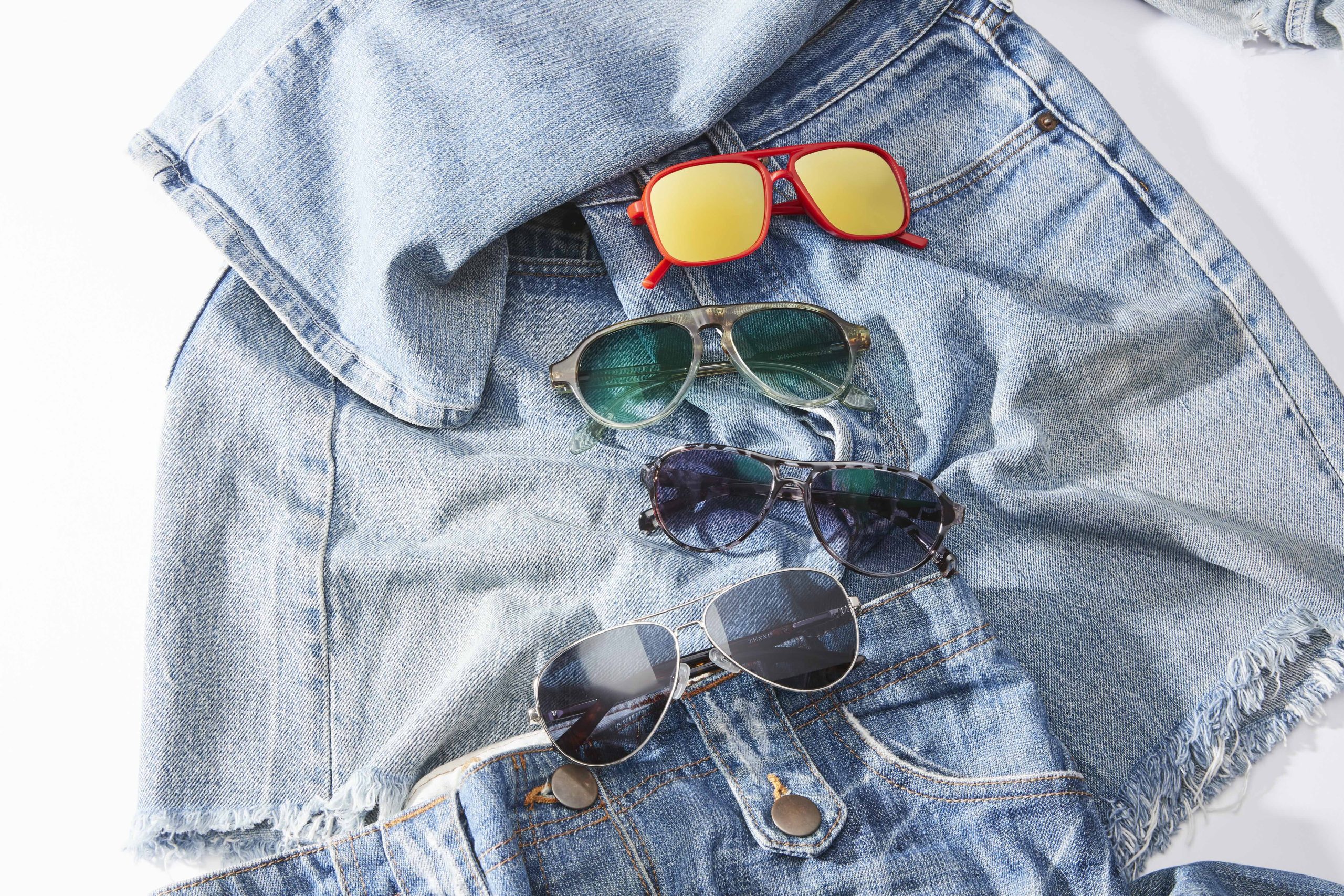 Four pairs of sunglasses on denim clothing, including red-framed, green-lensed, dark-lensed, and aviator styles.
