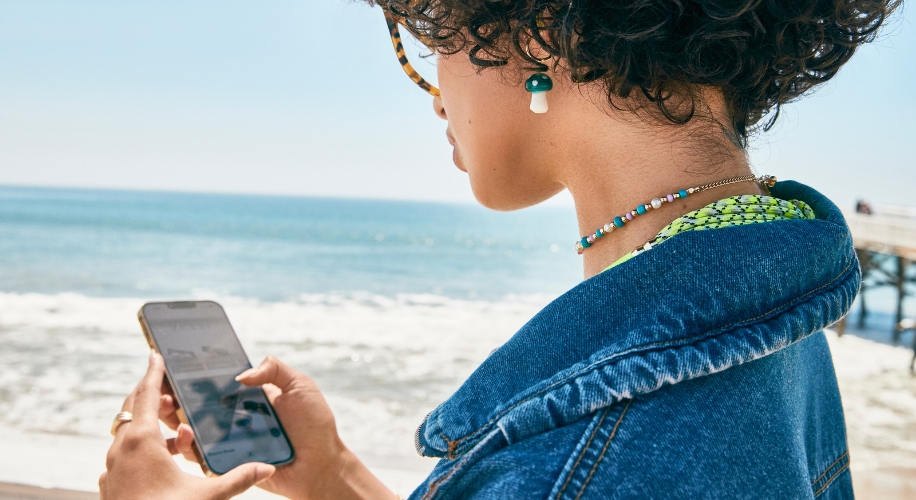 Person holding a smartphone near the beach, wearing a denim jacket, beaded necklace, and mushroom earring.