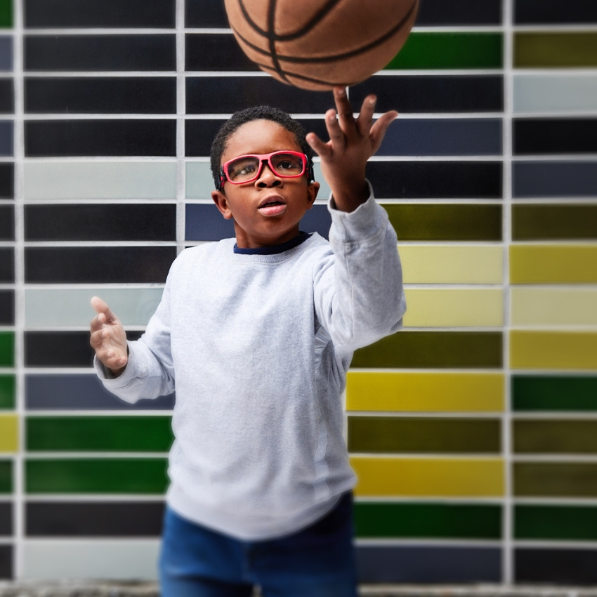 Child wearing red sports glasses, tossing a basketball while standing in front of a multicolored tile wall.