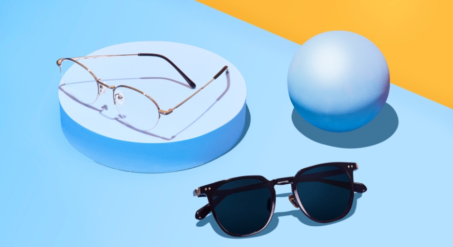 Round glasses with a gold frame and black sunglasses with a solid black frame on a blue circular platform.