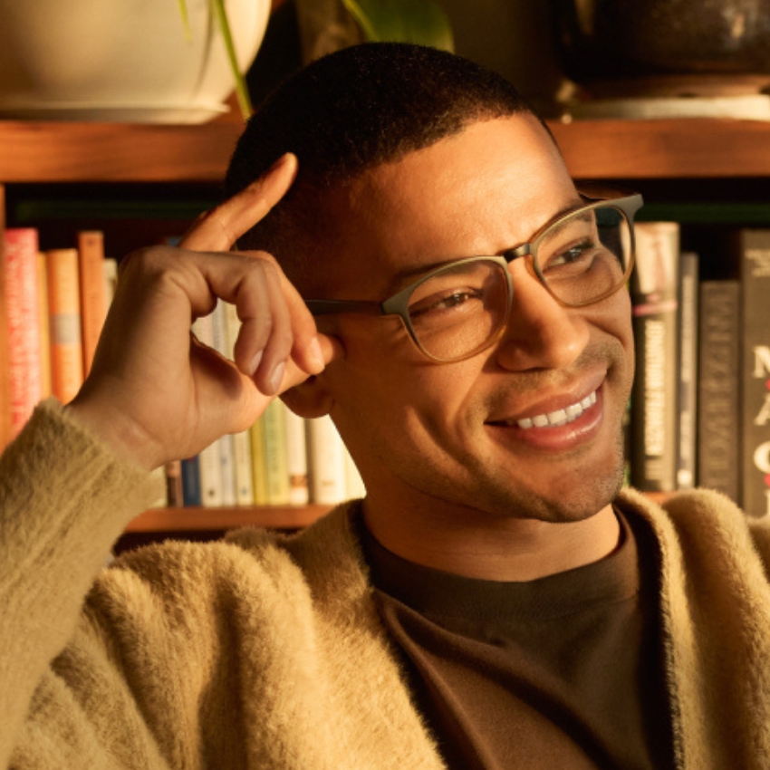 Man wearing dark-rimmed glasses and a beige sweater, smiling with his hand touching his temple.