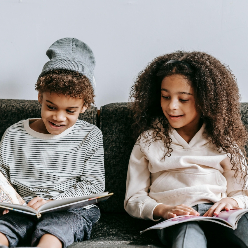 Two children sitting on a dark couch, both reading books and smiling.