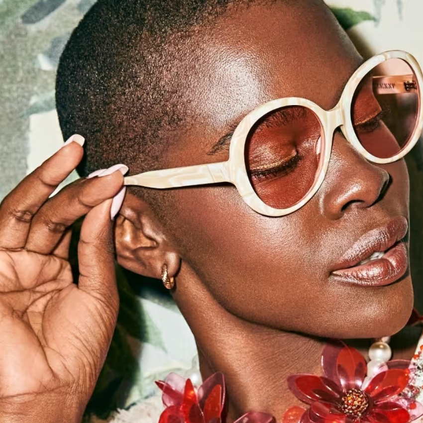 Woman wearing large round beige sunglasses with brown lenses and floral necklace. "DKNY" text visible on lens.