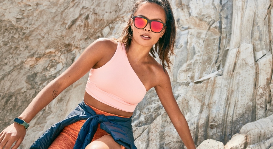 Woman wearing a pink one-shoulder sports top and red mirrored sunglasses. She has a blue jacket tied around her waist.