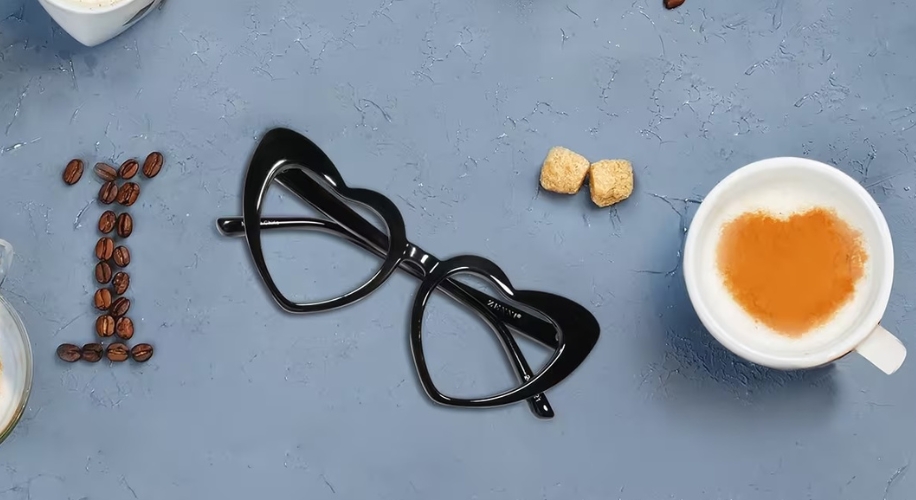 Black cat-eye glasses next to coffee cup with heart-shaped foam and coffee beans spelling &quot;I&quot;.