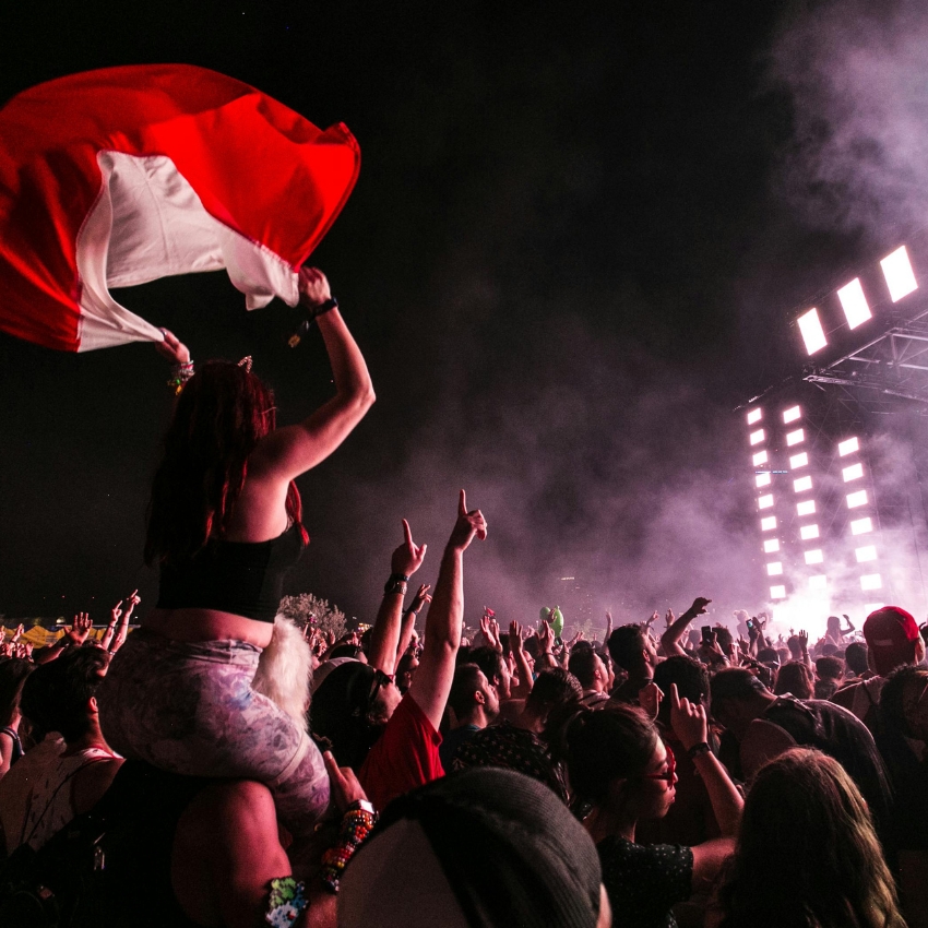 Person with a Peruvian flag on shoulders at a concert, surrounded by a crowd with raised arms and illuminated stage.