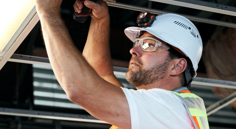 Construction worker with safety vest, helmet, and goggles using a drill on a metal frame.