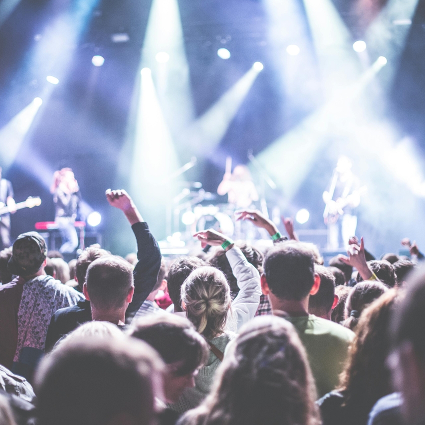Concert crowd with raised hands facing a stage, bright stage lights and band performing in the background.