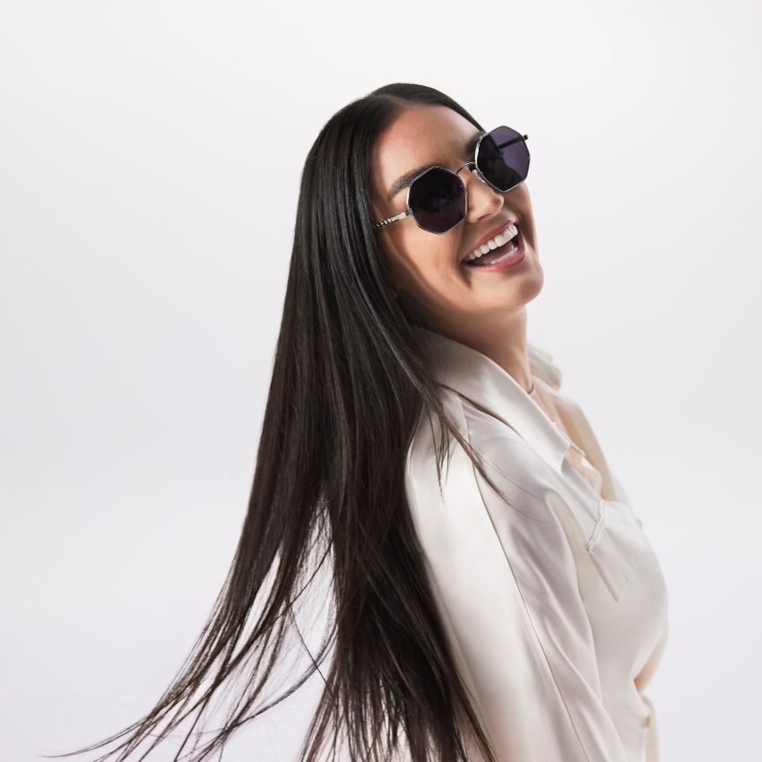 Woman wearing black round sunglasses and a white satin blouse, smiling and looking to the side.