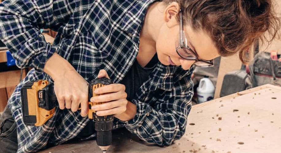 Person using a cordless drill on a wooden surface while wearing safety glasses and a plaid shirt.