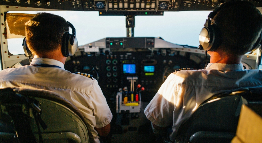 Two pilots in white uniforms and headsets seated in an airplane cockpit filled with instruments.