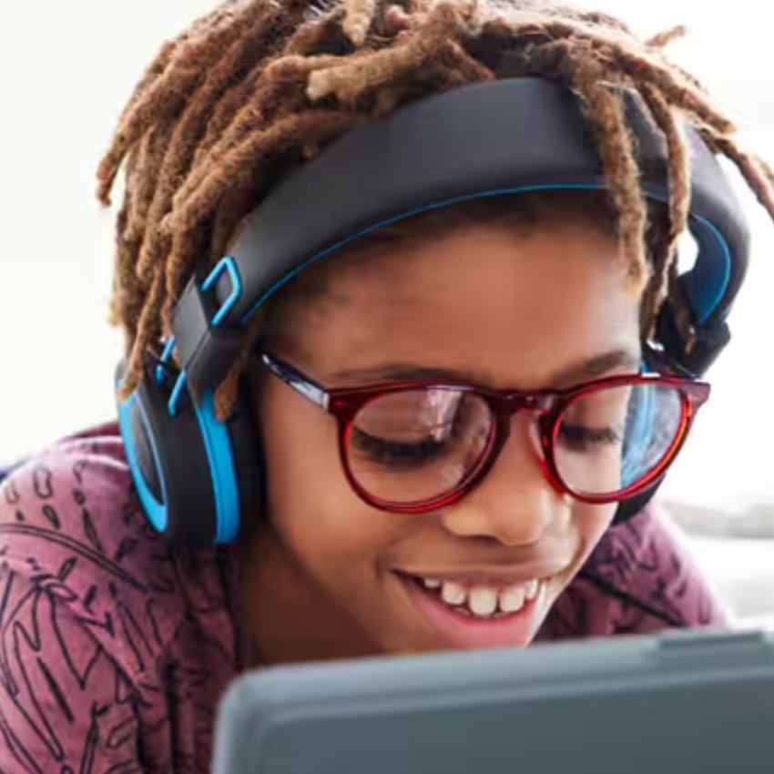 Child wearing red glasses and blue over-ear headphones, smiling while looking at a tablet.