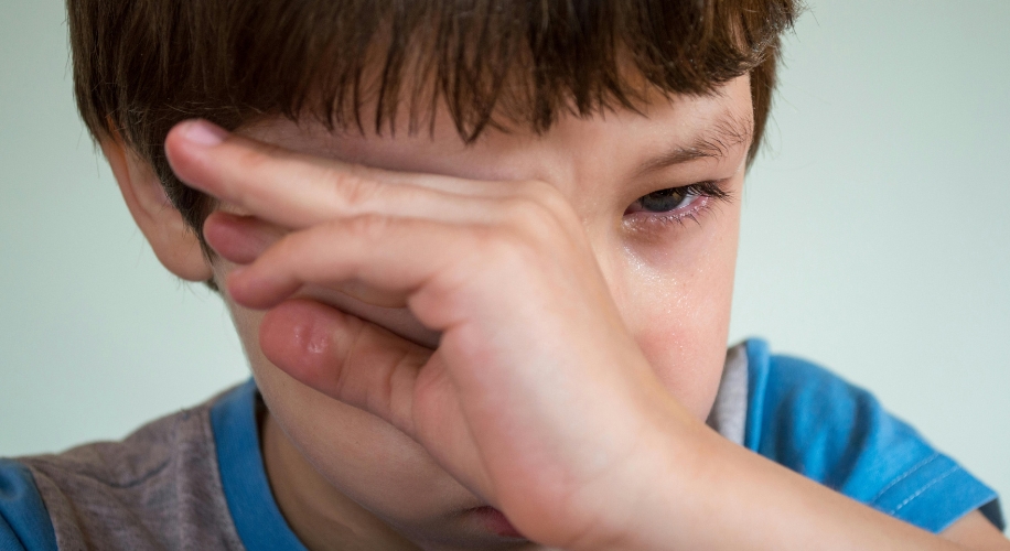 A child with brown hair rubbing their eye, wearing a blue and gray shirt.