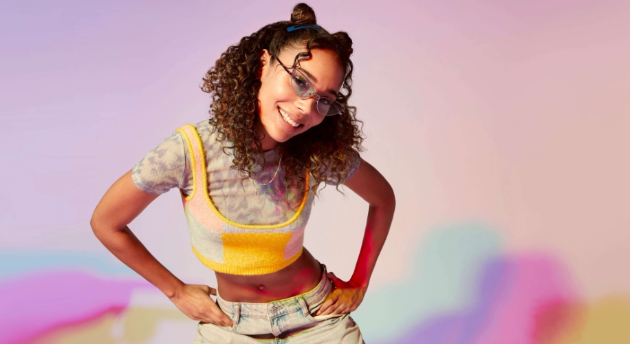 Woman wearing glasses, a yellow cropped top over a grey shirt, and light-colored jeans, hands on hips.