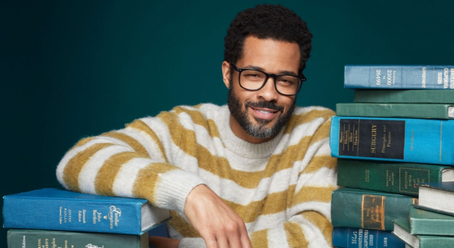 Man in glasses wearing a striped sweater, surrounded by stacks of books.