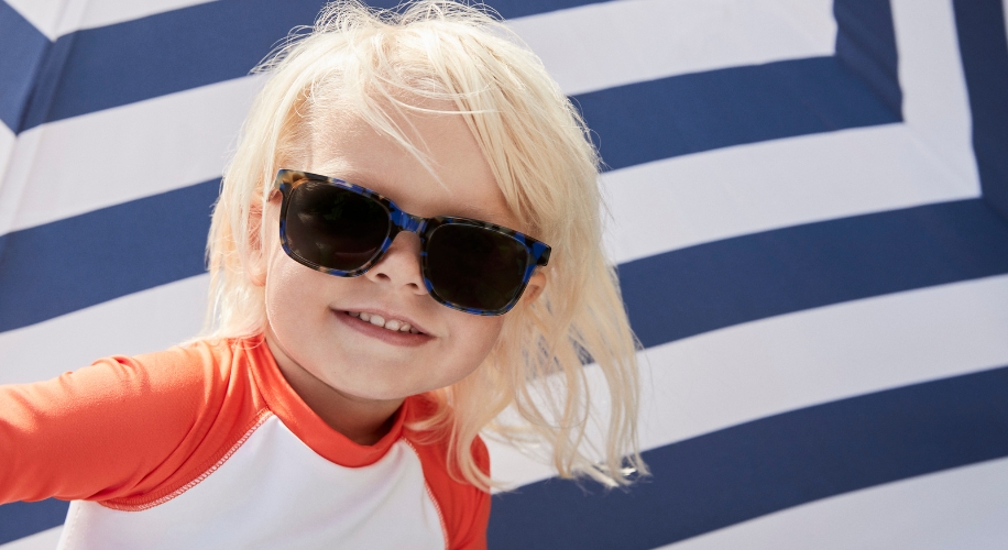 Child wearing tortoiseshell sunglasses and a red and white rash guard, smiling in front of a striped backdrop.