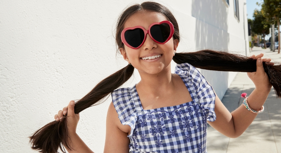 Child smiling, wearing heart-shaped sunglasses and a blue checkered dress, holding her pigtails.