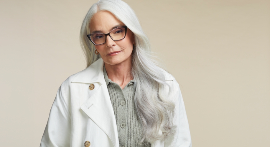 Senior woman with long silver hair models a white trench coat and glasses.
