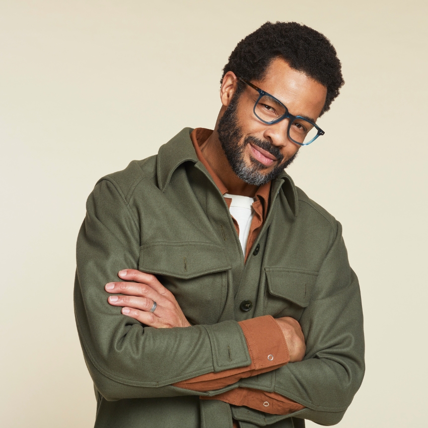 Man wearing blue-framed glasses, an olive green button-up shirt jacket, and an orange-brown undershirt.