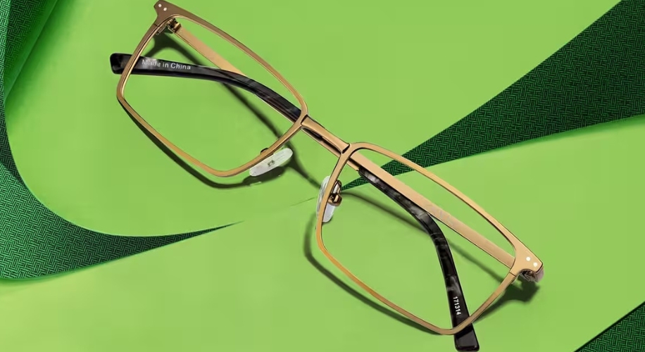 Gold rectangular eyeglasses on a green surface. Text on the temples: &quot;Made in China&quot; and &quot;ITALI&quot;.