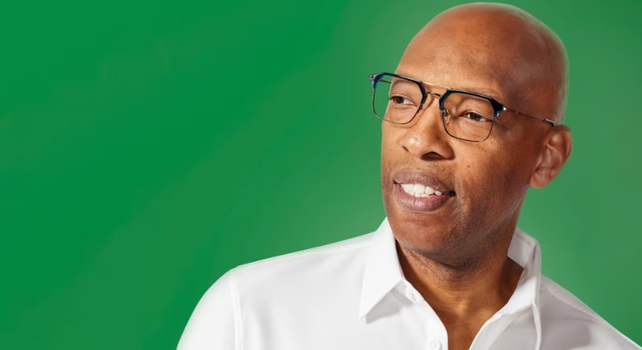 Bald man wearing glasses and a white collared shirt against a green background.