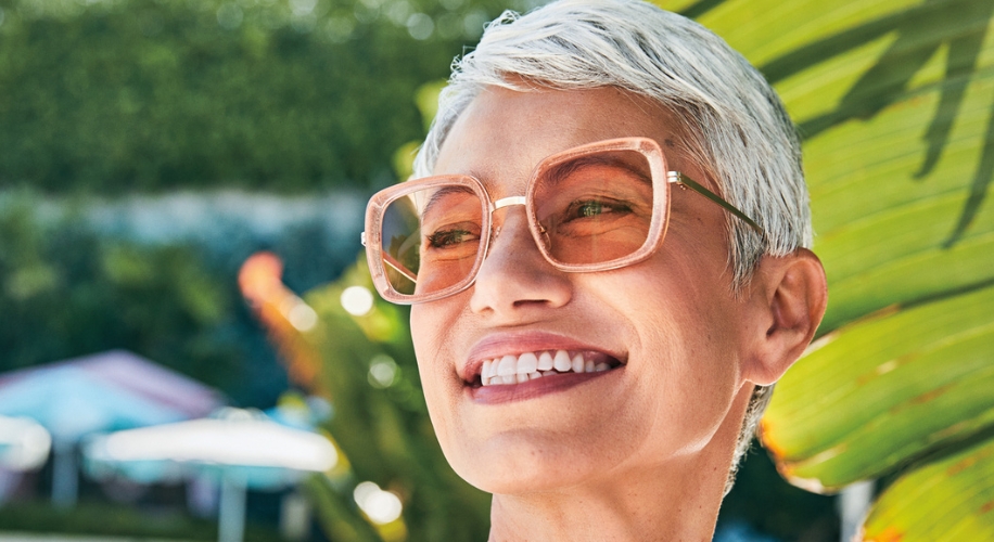 Woman wearing large square pink sunglasses with a gold bridge and arms, smiling outdoors.