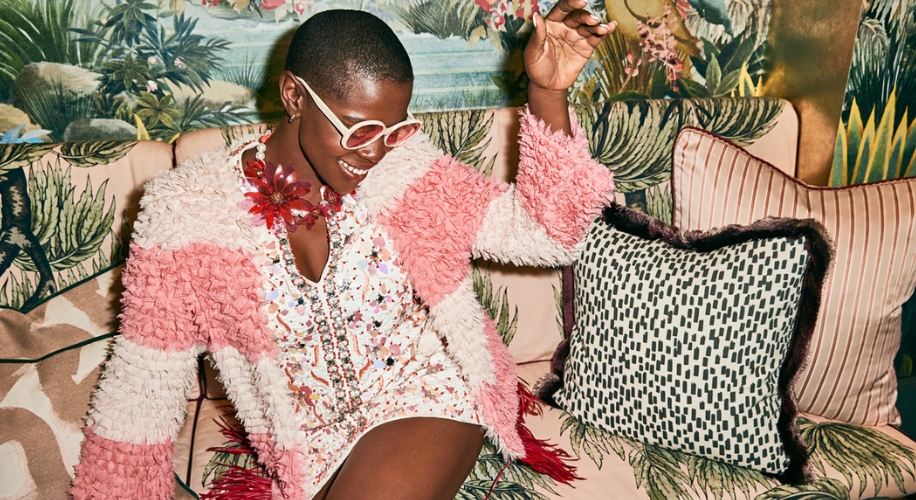 Woman wearing sunglasses, a pink and white textured sweater, beaded dress, floral necklace, seated on a sofa.
