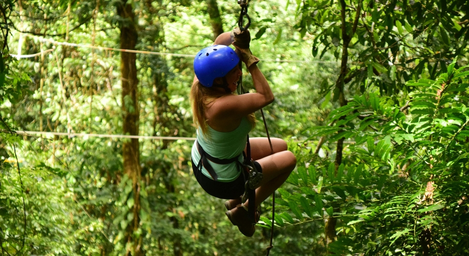 Person zip lining through a lush green forest, wearing a blue helmet and green tank top.