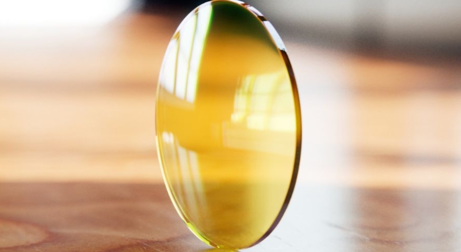 Yellow-tinted optical lens with reflective surface standing upright on a wooden surface.