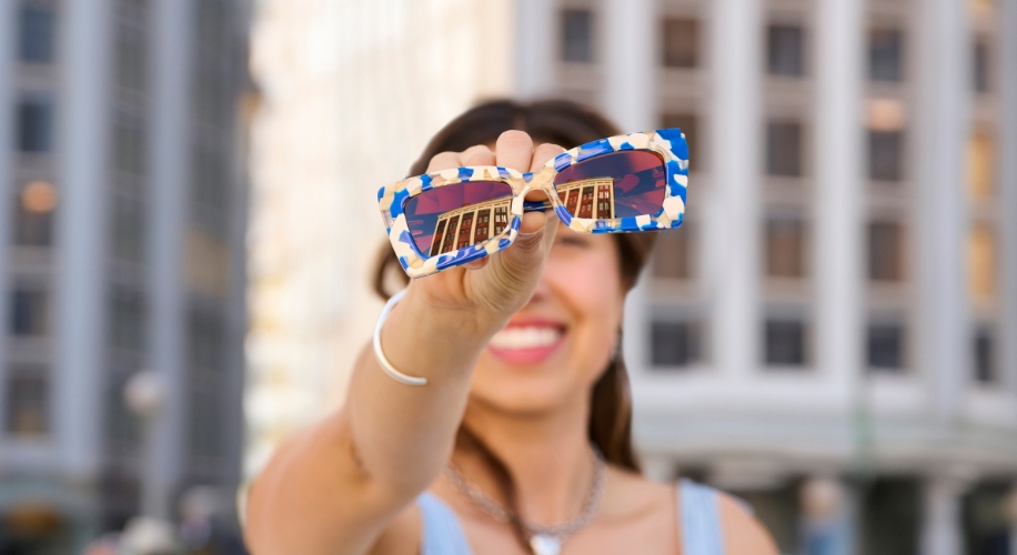 Woman holding blue and white patterned sunglasses with a mirrored reflection of a building.