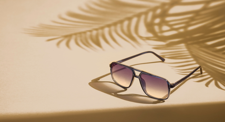Black-framed sunglasses with gradient lenses on a beige surface with palm leaf shadows.