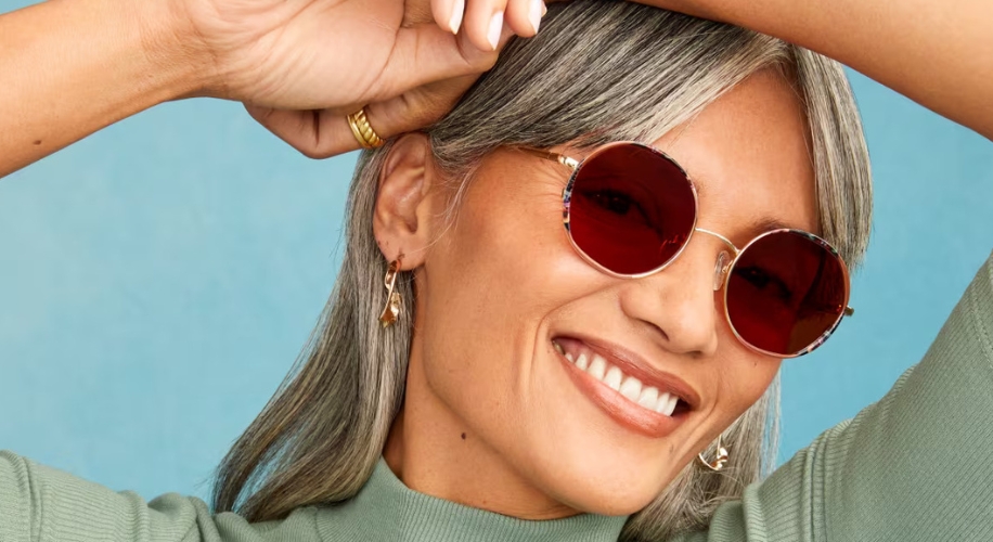 Woman wearing round, red-tinted sunglasses and gold earrings, smiling.