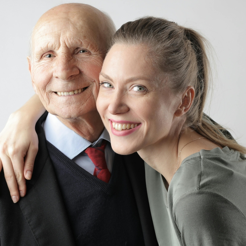 Smiling elderly man in a suit with a red tie and a younger woman embrace and pose for the camera.