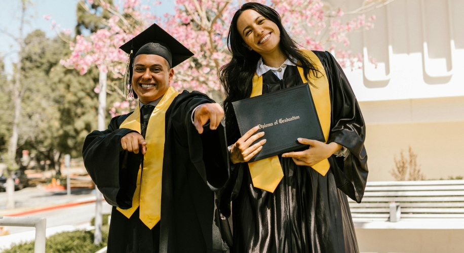 Graduates in caps and gowns, one holding a diploma that reads &quot;Diploma of Graduation.&quot;