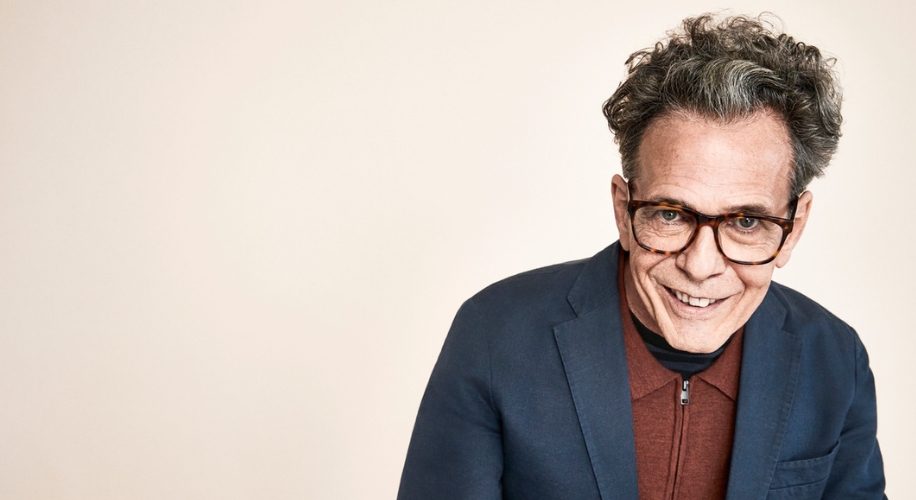 Smiling individual wearing a dark blue blazer over a brown zip-up top and glasses.