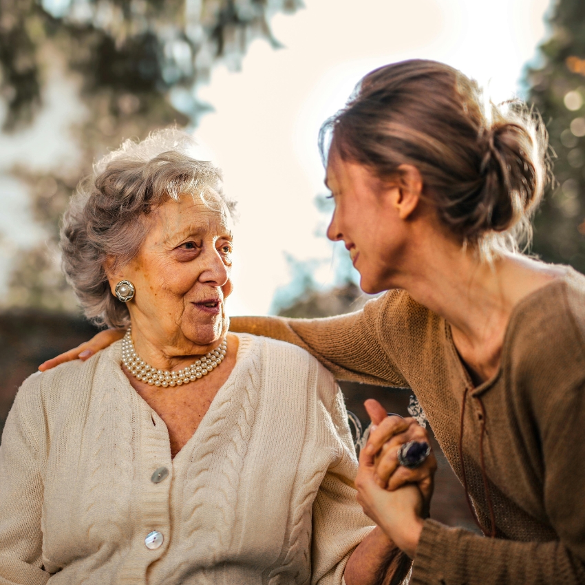 Elderly woman in a white cardigan holding hands with a younger woman, both smiling warmly.