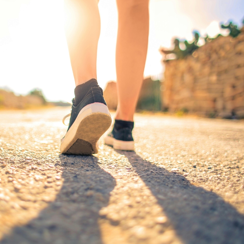 Person walking on a sunlit path wearing black athletic shoes with white soles.