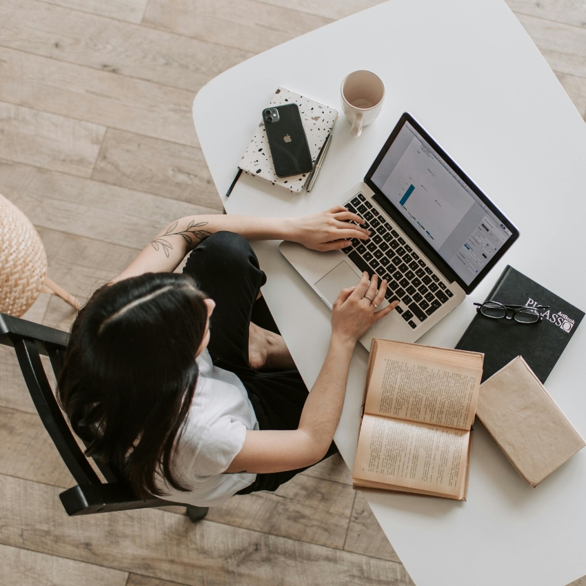 Person using a laptop at a white table, surrounded by books, a notebook, phone, pen, and a cup.