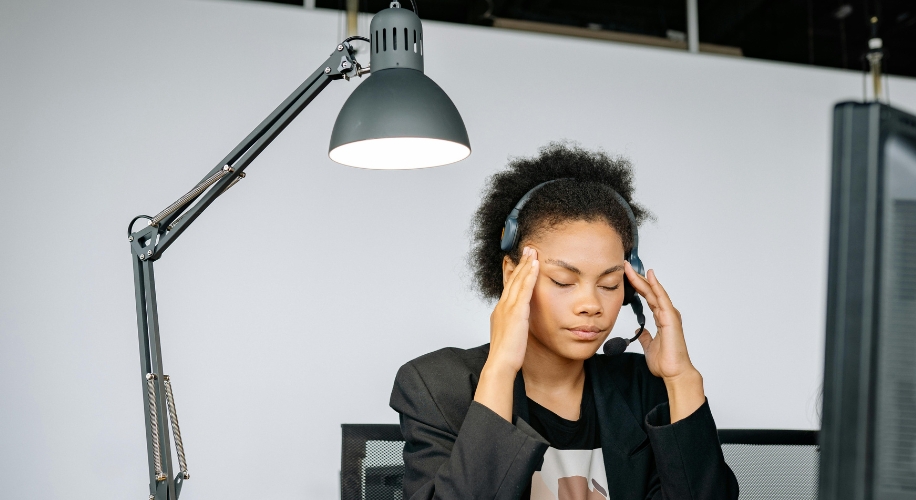 Person wearing a headset, sitting under a desk lamp, massaging temples and appearing stressed.