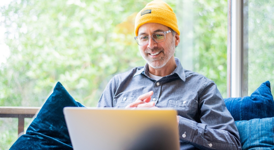 Person wearing a yellow beanie, glasses, and a gray shirt, working on a laptop on a couch.
