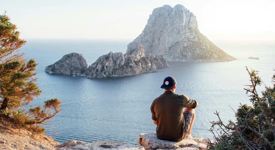 Person wearing a cap sitting on a rock and overlooking the ocean with a large rock island in the distance.
