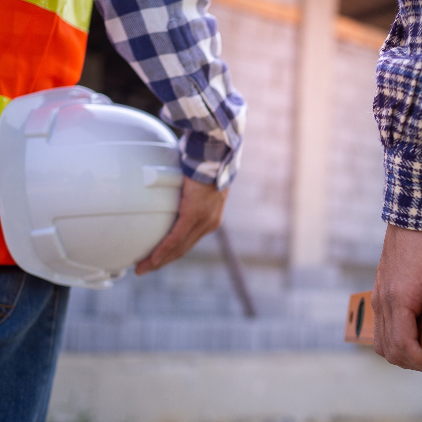 Construction worker holding a white hard hat, wearing a checkered shirt and safety vest.