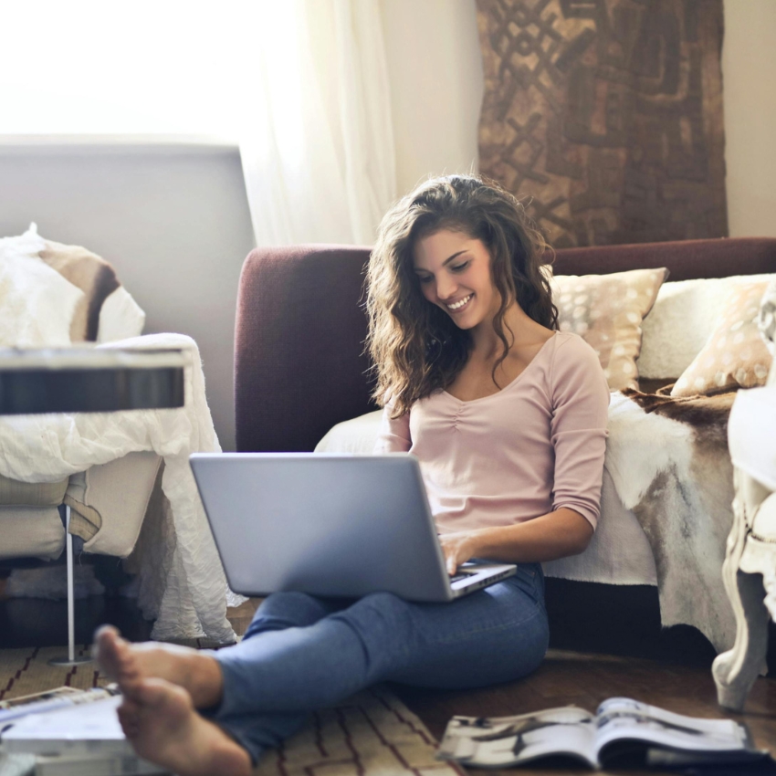 Smiling woman using a silver laptop on the floor; an open magazine lies beside her.