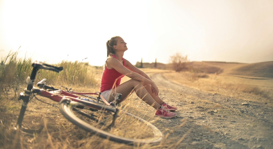 Woman in athletic wear resting on the ground beside a bicycle on a dirt path.