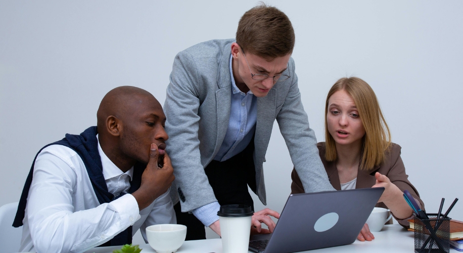 Three people gathered around a laptop, collaborating on work-related tasks. A paper coffee cup beside them.