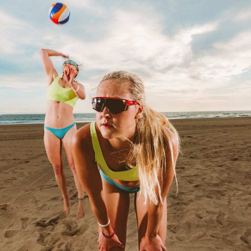 Two women playing beach volleyball, one wearing sunglasses and a neon green swimsuit preparing to return the ball, the other in the background serving the ball.