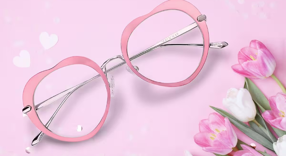 Pink heart-shaped eyeglasses with metal frames next to pink and white tulips on a pink background.