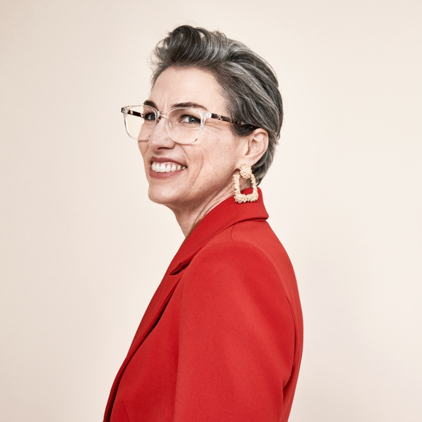 Gray-haired woman smiling in clear glasses, chunky earrings, and red blazer.