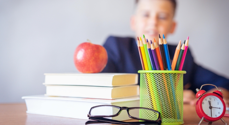 Colorful pencils in a green holder, stacked books with an apple, black eyeglasses, and a red alarm clock.